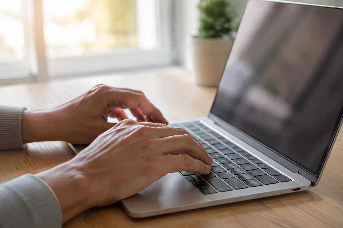 Mains posées sur un clavier d&rsquo;ordinateur portable devant une fenêtre, lumière naturelle du matin sur un bureau clair